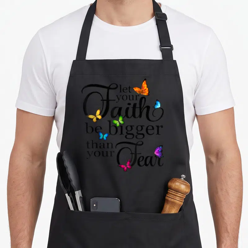 Bigger butterfly apron - Stacked black text let your Faith be bigger than your Fear surrounded by seven orange blue — shown on a black cotton canvas apron against a white background