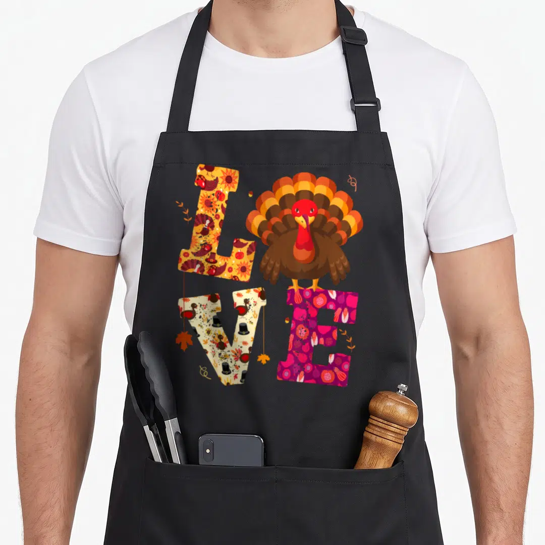 Teacher fall aprons - Cartoon turkey stands on purple E in LOVE filled with harvest patterns in orange, brown — shown on a black cotton canvas apron against a white background