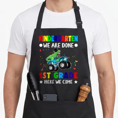 Grade come and get it apron - Dinosaur in cap on blue monster truck with rainbow text reading kindergarten done 1st — shown on a black cotton canvas apron against a white background