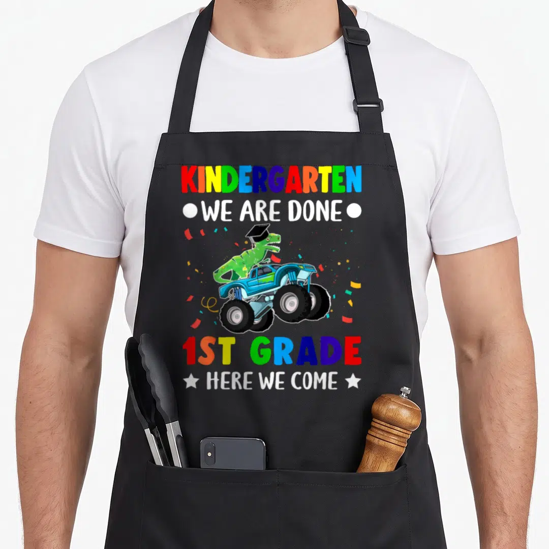 Grade come and get it apron - Dinosaur in cap on blue monster truck with rainbow text reading kindergarten done 1st — shown on a black cotton canvas apron against a white background
