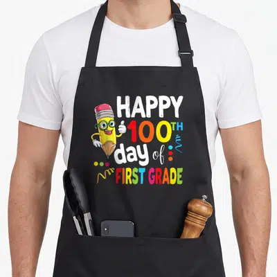 Retro 100th teacher apron - Cartoon pencil with glasses next to Happy 100th Day of First Grade text in multiple colors — shown on a black cotton canvas apron against a white background