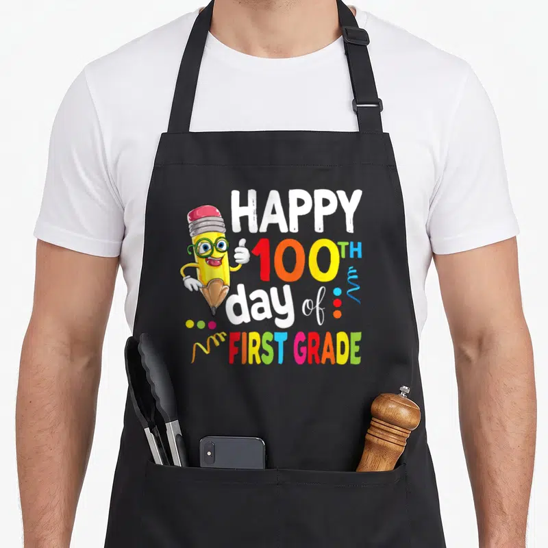 Retro 100th teacher apron - Cartoon pencil with glasses next to Happy 100th Day of First Grade text in multiple colors — shown on a black cotton canvas apron against a white background