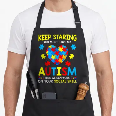 Staring eve apron - Sarcastic autism text with multicolored puzzle heart and scattered pieces — shown on a black cotton canvas apron against a white background