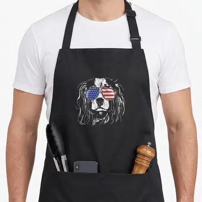 Flag king apron - White spaniel head wearing sunglasses with blue stars and red white stripes lenses — shown on a black cotton canvas apron against a white background