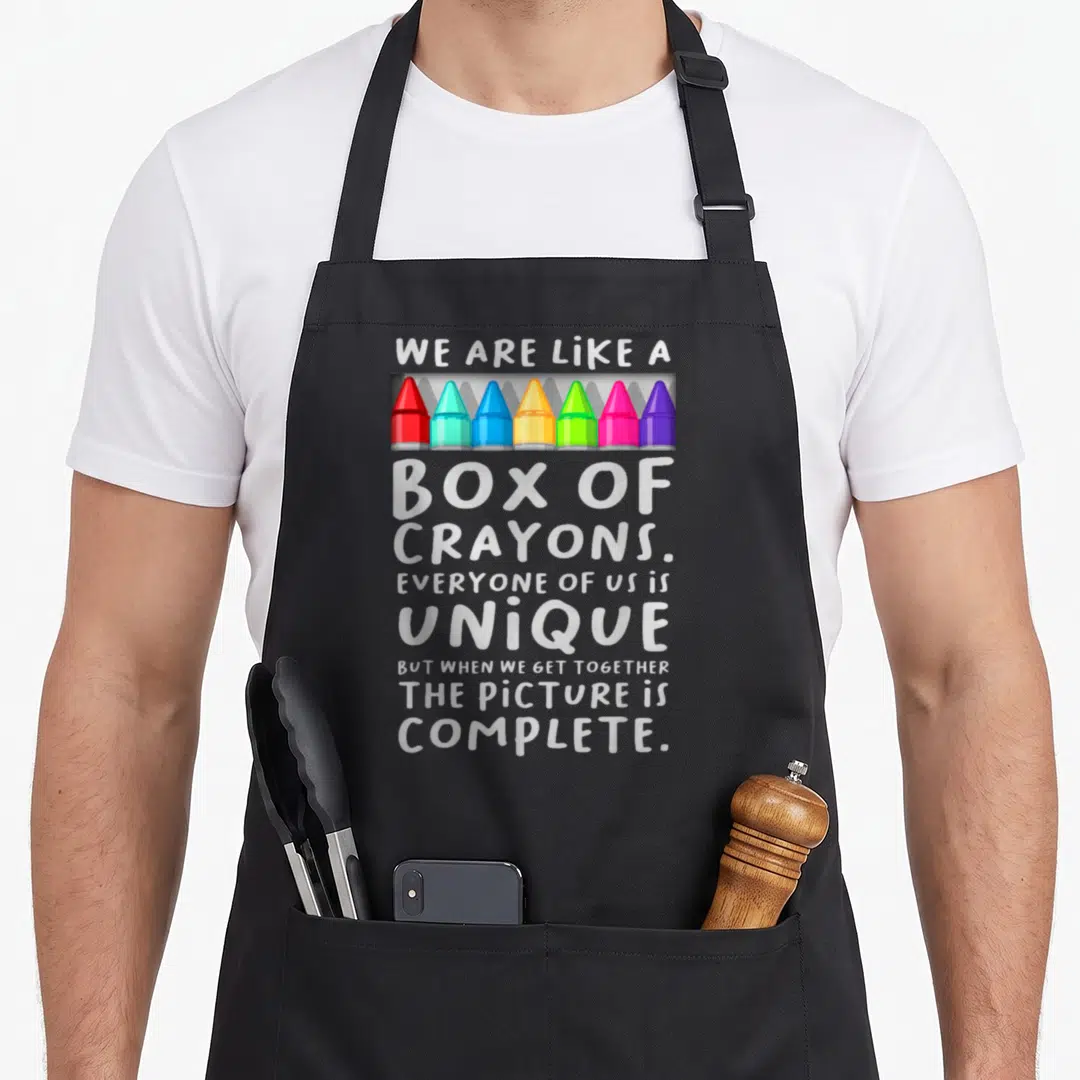 School box teacher apron - Seven colorful crayons in grey box with stacked white text about uniqueness — shown on a black cotton canvas apron against a white background