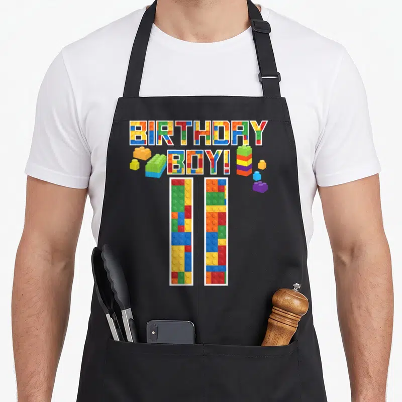 11th building aprons - BIRTHDAY BOY! and 11 text filled with red blue yellow and green building block patterns — shown on a black cotton canvas apron against a white background