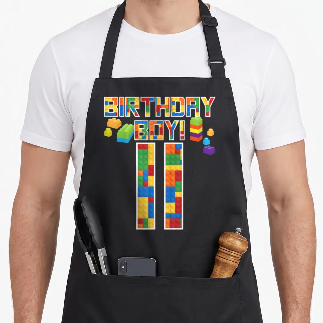 11th building aprons - BIRTHDAY BOY! and 11 text filled with red blue yellow and green building block patterns — shown on a black cotton canvas apron against a white background