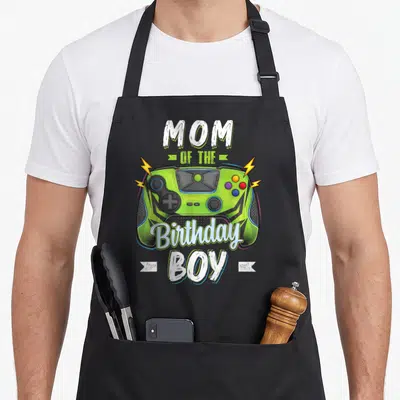 Gamer family aprons - Lime green game controller with white text reading Mom of the Birthday Boy surrounded by yellow — shown on a black cotton canvas apron against a white background