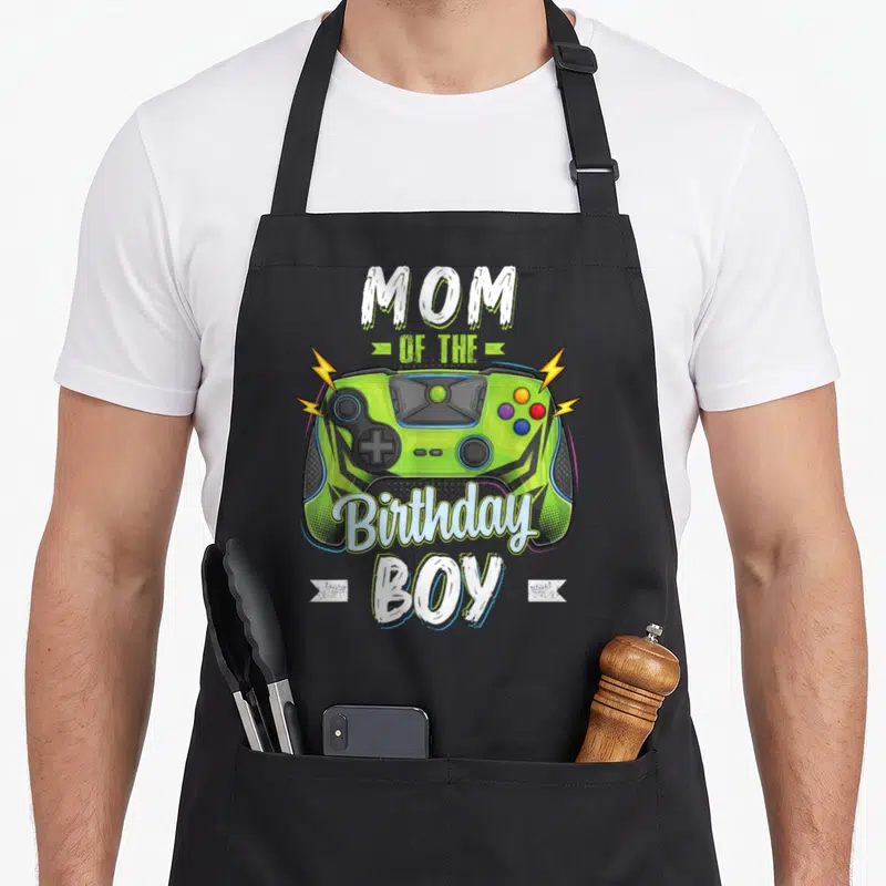 Gamer family aprons - Lime green game controller with white text reading Mom of the Birthday Boy surrounded by yellow — shown on a black cotton canvas apron against a white background