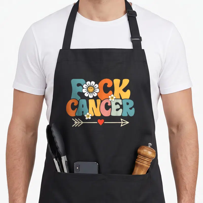 Groovy breast cancer awareness aprons - FUCK CANCER text in blue orange pink yellow with smiley daisies and heart arrow — shown on a black cotton canvas apron against a white background