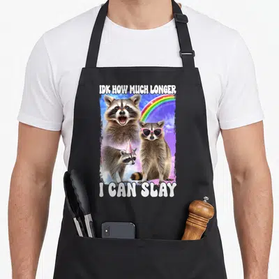 Much meme apron - Three raccoons, rainbow, pink sunglasses, party hat, white text on purple cloudy background — shown on a black cotton canvas apron against a white background