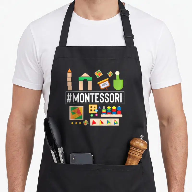 Studying montessori teacher apron - Cartoon educational toys in red yellow blue green beige surrounding a white — shown on a black cotton canvas apron against a white background