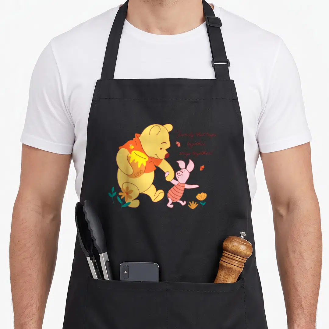 Piglet family aprons - Winnie the Pooh and Piglet walking with honey pot and text Family that trips together stays — shown on a black cotton canvas apron against a white background