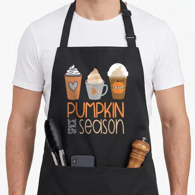 Spice fall aprons - Three cartoon coffee cups above stacked PUMPKIN SPICE season text in orange and beige — shown on a black cotton canvas apron against a white background