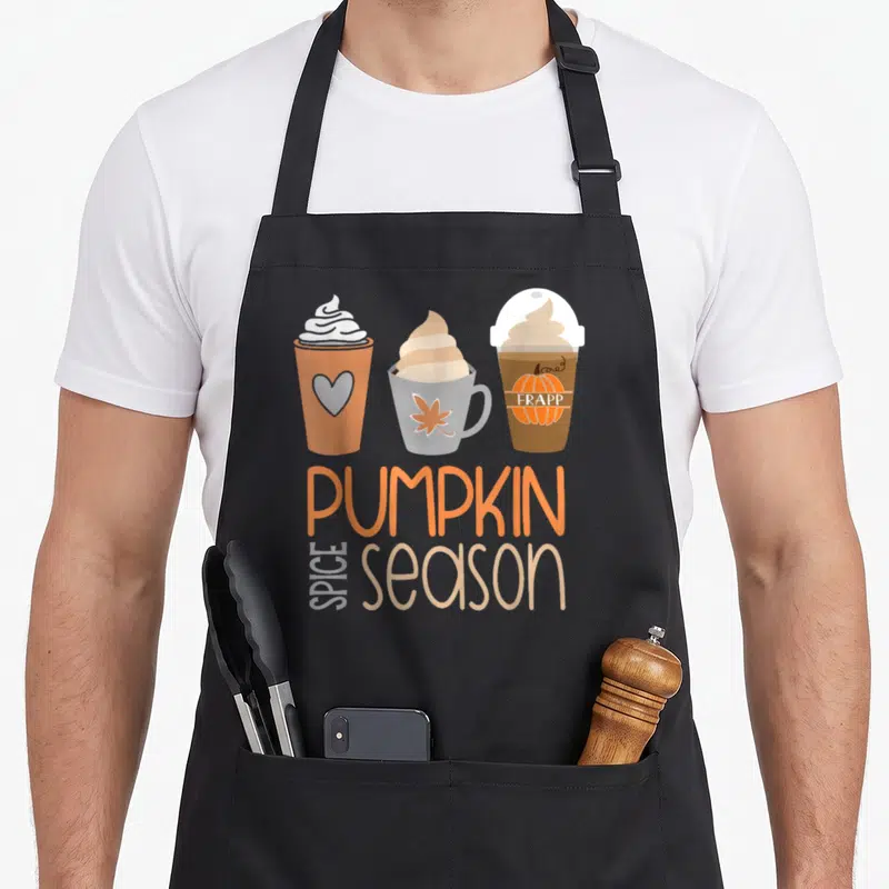 Spice fall aprons - Three cartoon coffee cups above stacked PUMPKIN SPICE season text in orange and beige — shown on a black cotton canvas apron against a white background