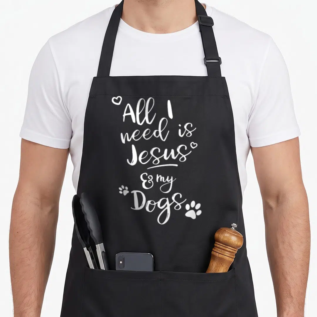 Jesus dog apron - White stacked text All I need is Jesus & my Dogs with hearts and paw prints — shown on a black cotton canvas apron against a white background