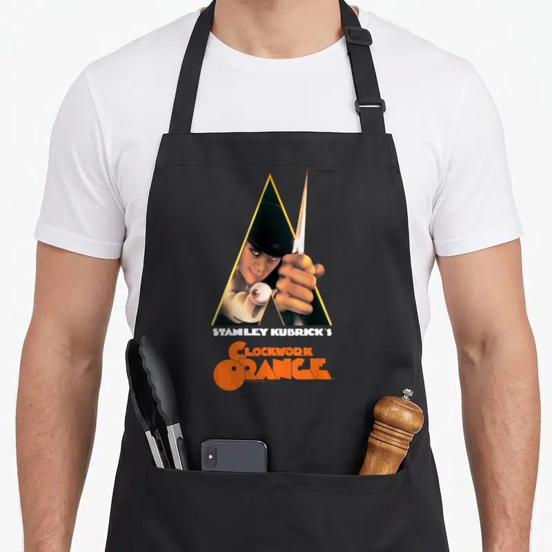 Clockwork novelty apron - Man holding knife in yellow triangle with Stanley Kubrick's Clockwork Orange text — shown on a black cotton canvas apron against a white background