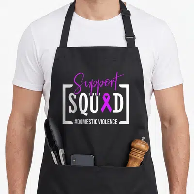 Squad domestic aprons - Purple script Support text over white SQUAD bracketed text with purple ribbon replacing — shown on a black cotton canvas apron against a white background