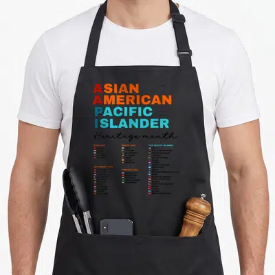 Aapi pacific asian apron - ASIAN AMERICAN PACIFIC ISLANDER text with five regional country lists and flags in red — shown on a black cotton canvas apron against a white background