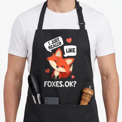 Funny animal fox aprons - Cartoon fox surrounded by hearts and speech bubbles reading I Just Really Like Foxes OK — shown on a black cotton canvas apron against a white background