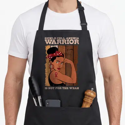 Cell blood apron - Cartoon woman with red polka dot headscarf and ribbon flexing arm in front of brown wood planks — shown on a black cotton canvas apron against a white background