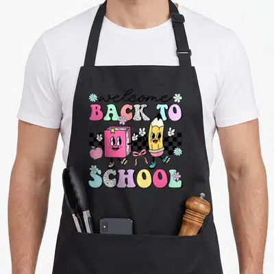 Funny student art teacher apron - Cartoon book and pencil walking over checkerboard with welcome back to school text — shown on a black cotton canvas apron against a white background