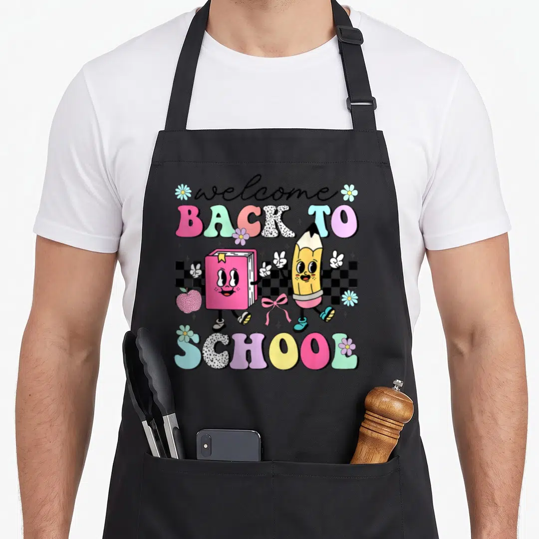 Funny student art teacher apron - Cartoon book and pencil walking over checkerboard with welcome back to school text — shown on a black cotton canvas apron against a white background