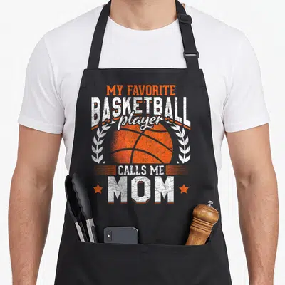 Call basketball player apron - Orange basketball graphic with white text and two stars flanking the word mom — shown on a black cotton canvas apron against a white background