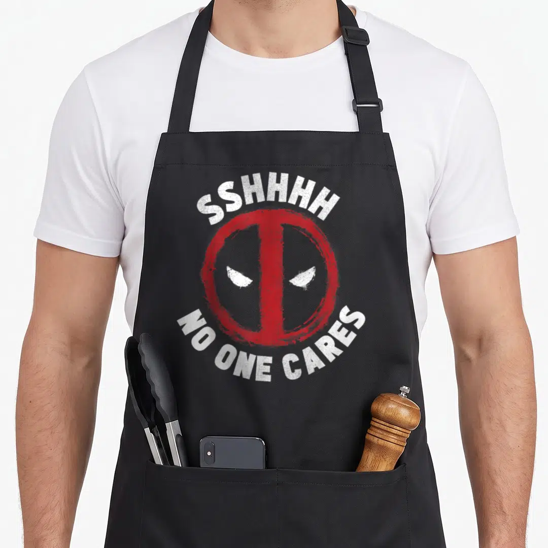 Funny sshhhh care apron - Red circle with white Deadpool eyes and arched text reading SSHHHH NO ONE CARES in distressed — shown on a black cotton canvas apron against a white background