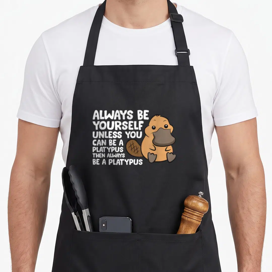 Alway platypus mom apron - Cartoon brown platypus sitting beside white stacked text reading Always Be Yourself Unless — shown on a black cotton canvas apron against a white background