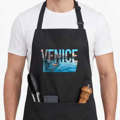 Venezia italy apron - Word VENICE masking a photo of a gondola, canal water, and buildings in blue and orange — shown on a black cotton canvas apron against a white background