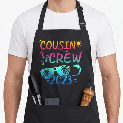 Cousin camp chef apron - COUSIN CREW 2023 text with sun, flip flops, sunglasses, and wave in orange pink teal blue — shown on a black cotton canvas apron against a white background
