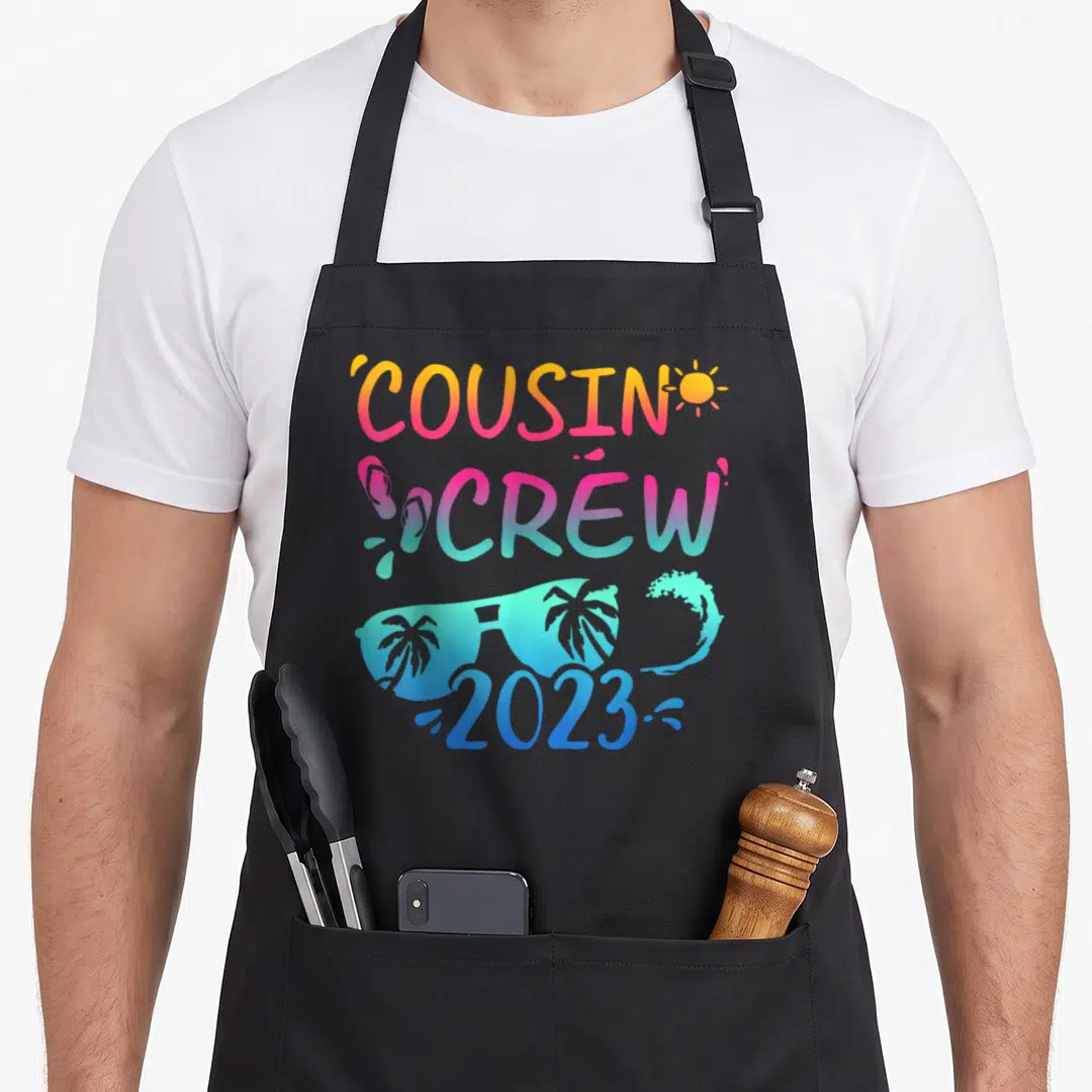 Cousin camp chef apron - COUSIN CREW 2023 text with sun, flip flops, sunglasses, and wave in orange pink teal blue — shown on a black cotton canvas apron against a white background