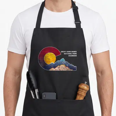 Sand themed aprons - Retro Great Sand Dunes National Park Colorado logo with sun and mountains in red yellow blue tan — shown on a black cotton canvas apron against a white background