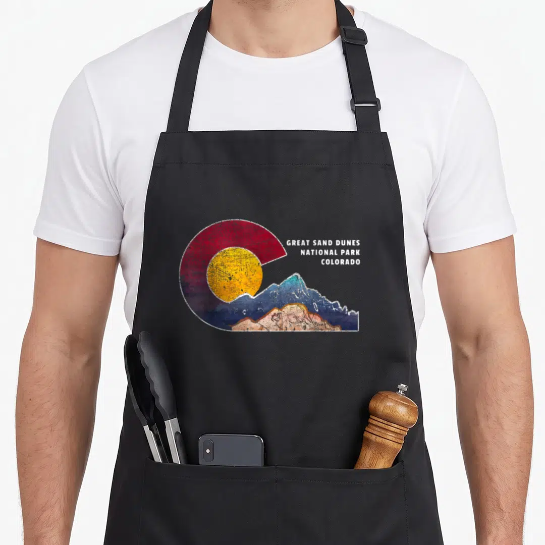 Sand themed aprons - Retro Great Sand Dunes National Park Colorado logo with sun and mountains in red yellow blue tan — shown on a black cotton canvas apron against a white background