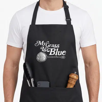 Blue banjo apron - White stacked text My Grass Is Blue above white banjo illustration distressed style — shown on a black cotton canvas apron against a white background