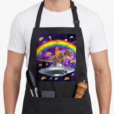 Taco kitty cat apron - Orange cat riding T-Rex on UFO with laser eyes, rainbow, tacos, and ice cream in space — shown on a black cotton canvas apron against a white background