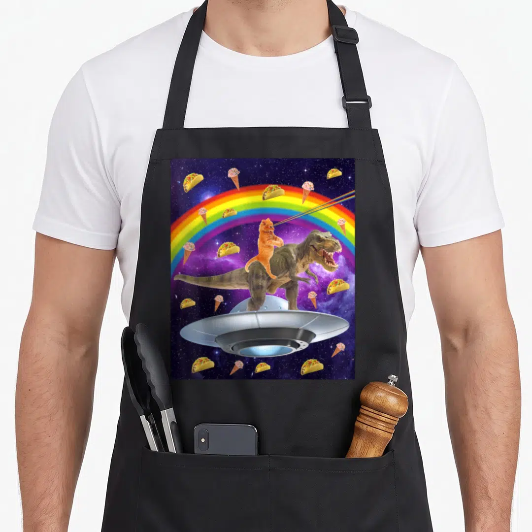 Taco kitty cat apron - Orange cat riding T-Rex on UFO with laser eyes, rainbow, tacos, and ice cream in space — shown on a black cotton canvas apron against a white background