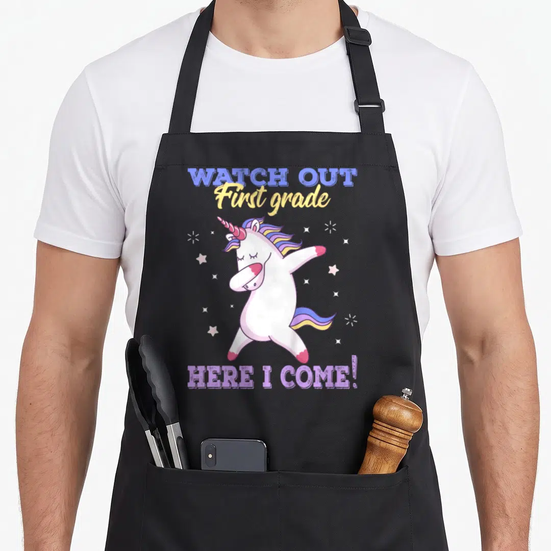 Watch grade apron - Dabbing unicorn with WATCH OUT First grade HERE I COME text and scattered stars in blue, yellow — shown on a black cotton canvas apron against a white background