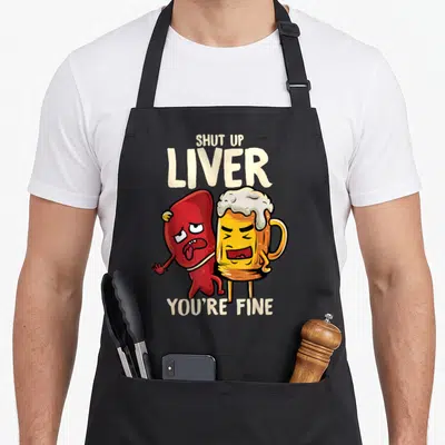 Shut beer apron - Cartoon red liver and yellow beer mug with Shut Up Liver You're Fine text — shown on a black cotton canvas apron against a white background