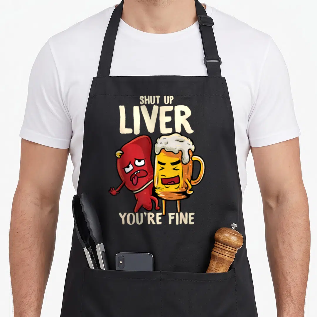 Shut beer apron - Cartoon red liver and yellow beer mug with Shut Up Liver You're Fine text — shown on a black cotton canvas apron against a white background