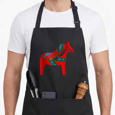 Dala horse swedish apron - Red dala horse with blue and green floral patterns facing right — shown on a black cotton canvas apron against a white background