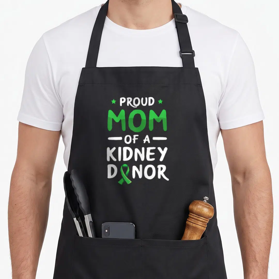 Kidney donor organ apron - Stacked white and green text reading PROUD MOM OF A KIDNEY DONOR with green ribbon in O — shown on a black cotton canvas apron against a white background