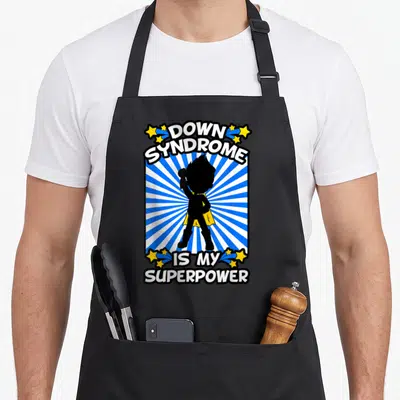 Syndrome superhero apron - DOWN SYNDROME IS MY SUPERPOWER text with caped child silhouette on blue sunburst and yellow — shown on a black cotton canvas apron against a white background