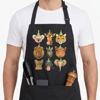 Wildlife zoo birthday apron - Nine cartoon animal heads in a grid wearing party hats and glasses — shown on a black cotton canvas apron against a white background