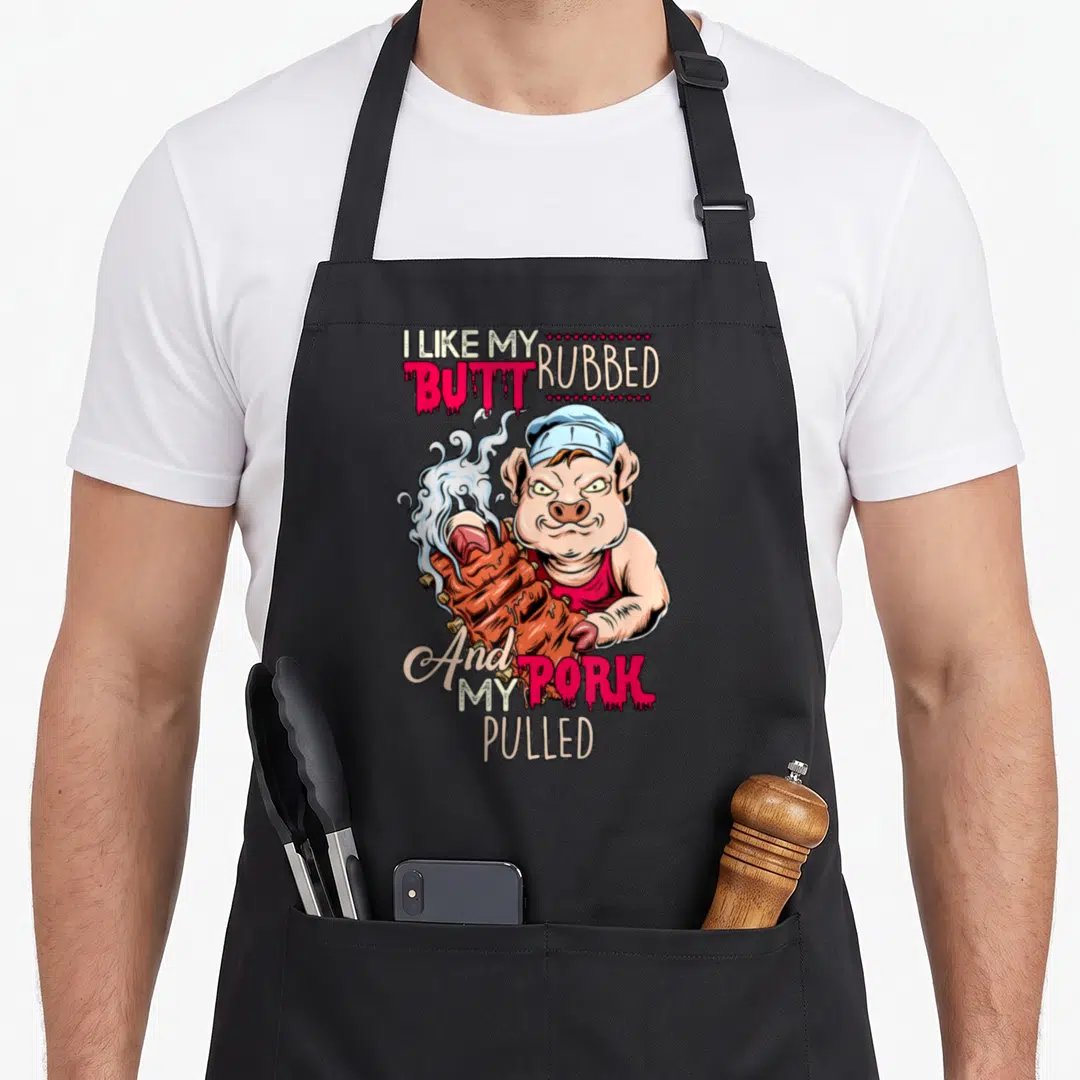 Butt grillmaster apron - Cartoon pig holding ribs with text I like my butt rubbed and my pork pulled — shown on a black cotton canvas apron against a white background