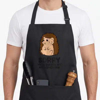 Sorry homework apron - Cartoon hedgehog sitting above text SORRY MY HEDGEHOG ATE MY HOMEWORK in brown and black — shown on a black cotton canvas apron against a white background