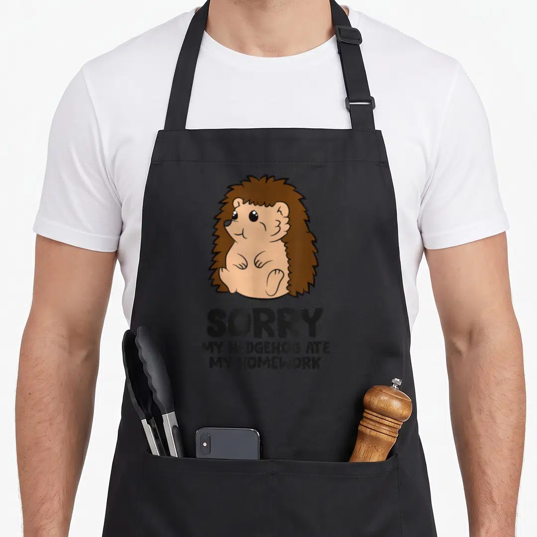 Sorry homework apron - Cartoon hedgehog sitting above text SORRY MY HEDGEHOG ATE MY HOMEWORK in brown and black — shown on a black cotton canvas apron against a white background