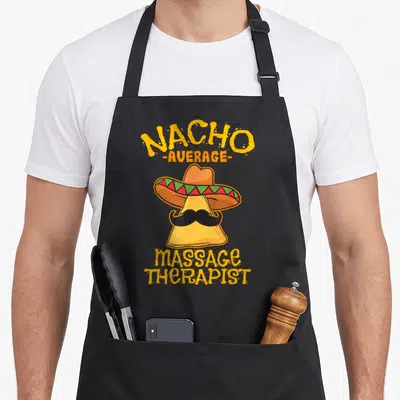 Nacho massage therapist apron - Cartoon nacho chip with sombrero and mustache, text NACHO -AVERAGE- massage THERAPIST — shown on a black cotton canvas apron against a white background