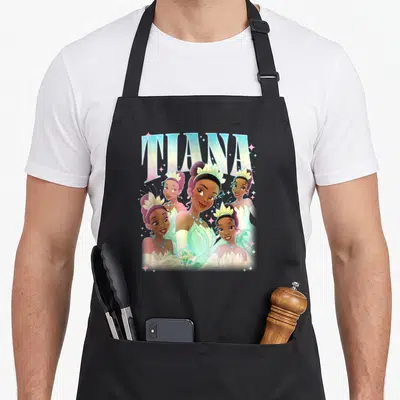 Beautiful princess apron - TIANA text above four overlapping cartoon depictions in blue, white, yellow, and brown — shown on a black cotton canvas apron against a white background